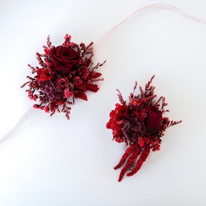 Puede incluir: Dos ramilletes florales para la muñeca con rosas de color rojo intenso y flores de color burdeos variadas. Cada ramillete está adornado con pequeñas flores secas y una delicada cinta rosa claro. El arreglo está sobre un fondo blanco.