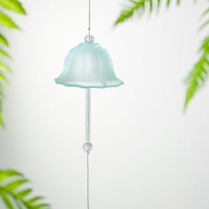 Pode incluir: Um delicado sino de vento de vidro azul claro com um design em forma de sino. O sino pende de um fio fino, com uma conta transparente no topo e na parte inferior. Folhas verdes de samambaia emolduram o sino contra um fundo branco.