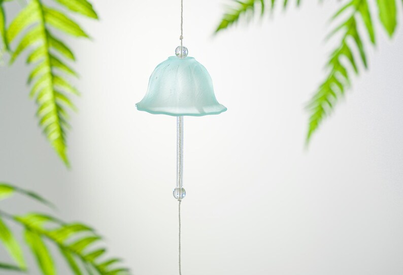 Pode incluir: Um delicado sino de vento de vidro azul claro com um design em forma de sino. O sino pende de um fio fino, com uma conta transparente no topo e na parte inferior. Folhas verdes de samambaia emolduram o sino contra um fundo branco.