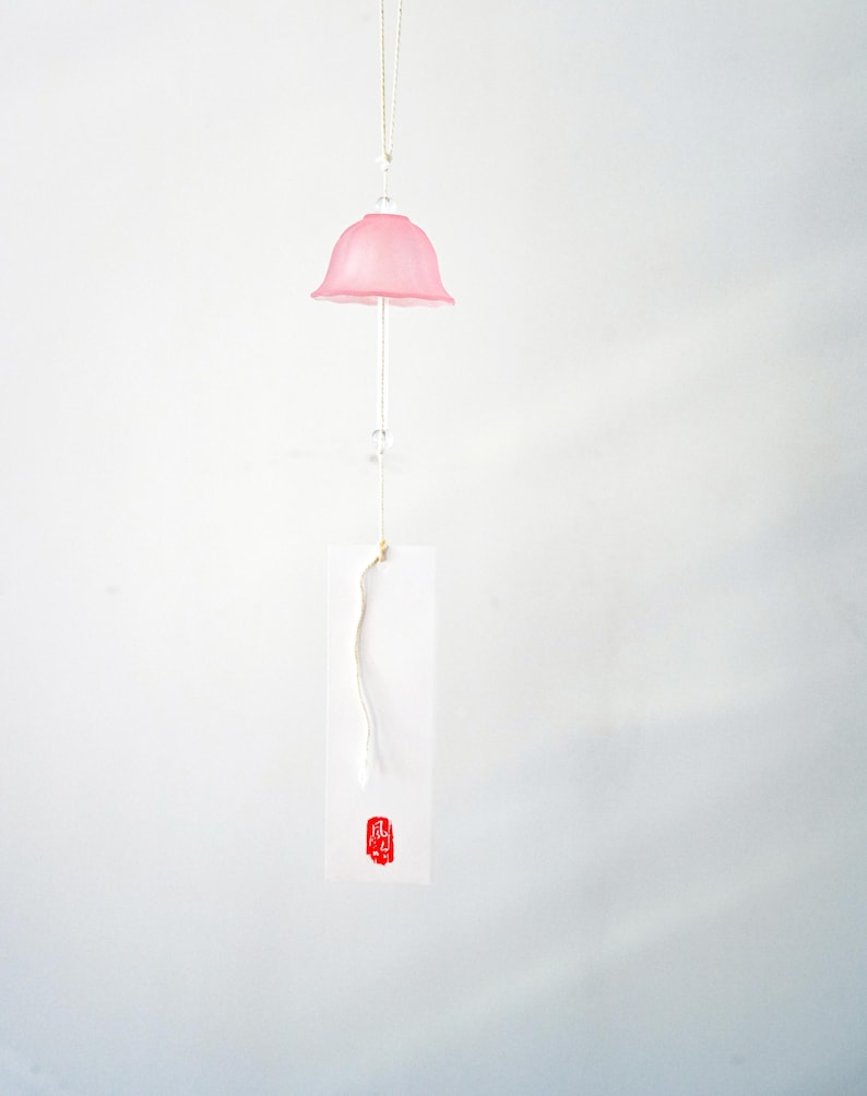 Pode incluir: Um sino de vento de vidro rosa com um topo em forma de sino, suspenso por um fio fino. Uma etiqueta de papel branco com um selo vermelho pende por baixo. O sino de vento est&aacute; contra um fundo branco.