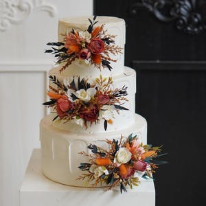 May include: A three-tiered white wedding cake decorated with cascading frosting and floral arrangements. The flowers are a mix of white, orange, and red.