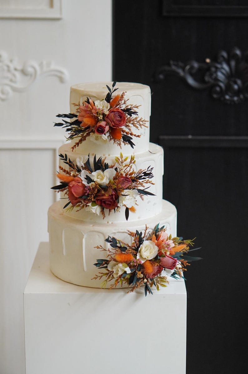 May include: A three-tiered white wedding cake decorated with cascading frosting and floral arrangements. The flowers are a mix of white, orange, and red.