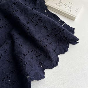 May include: A dark navy blue fabric featuring an intricate eyelet embroidery pattern with floral motifs and a scalloped edge. Next to the fabric is a white book with the text Rayon de Soleil printed on the cover in a black cursive font.