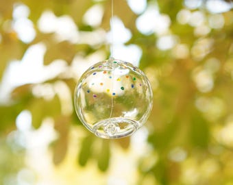 Könnte beinhalten: Ein klarer Glaswindspiel mit bunten Punkten, das an einer Schnur hängt, vor einem verschwommenen Hintergrund aus grünen Blättern.