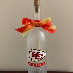 May include: A frosted glass wine bottle with a red and yellow bow, a white and red Kansas City Chiefs logo, and the text "CHIEFS".