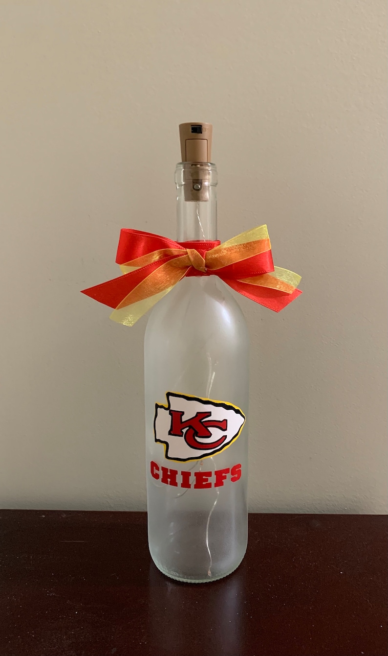 May include: A frosted glass wine bottle with a red and yellow bow, a white and red Kansas City Chiefs logo, and the text "CHIEFS".