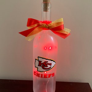May include: A clear glass bottle with a red and gold bow, a red and white Kansas City Chiefs logo, and red LED lights inside. The bottle is sitting on a brown surface.