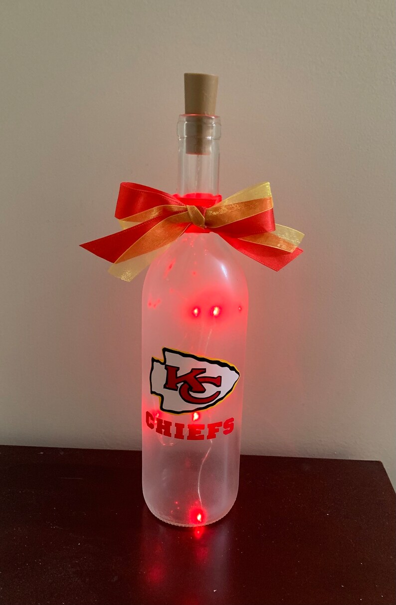 May include: A clear glass bottle with a red and gold bow, a red and white Kansas City Chiefs logo, and red LED lights inside. The bottle is sitting on a brown surface.