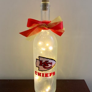 May include: A clear glass bottle with a cork top and a red and gold bow. The bottle is decorated with a white and red Kansas City Chiefs logo and has white fairy lights inside.