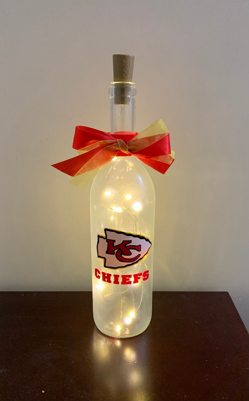 May include: A clear glass bottle with a cork top and a red and gold bow. The bottle is decorated with a white and red Kansas City Chiefs logo and has white fairy lights inside.