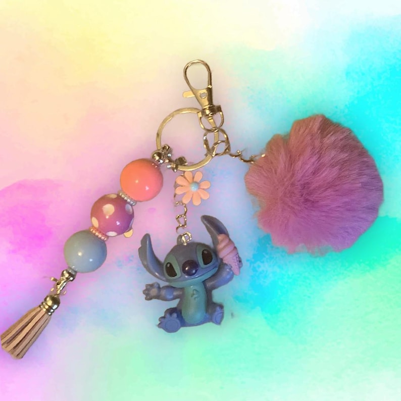 Puede incluir: Un llavero hecho a mano con una figura azul de Stitch sosteniendo un helado, un pomp&oacute;n rosa esponjoso y una borla de cuentas. El llavero incluye un mosquet&oacute;n y un dije de flor. Las cuentas son rosas, azules y naranjas.
