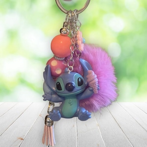 Puede incluir: Un llavero de Stitch azul y turquesa con un helado rosa, un pomp&oacute;n rosa y una borla rosa. El llavero tambi&eacute;n presenta una cuenta de lunares rosa y blanca y un dije de flor rosa.