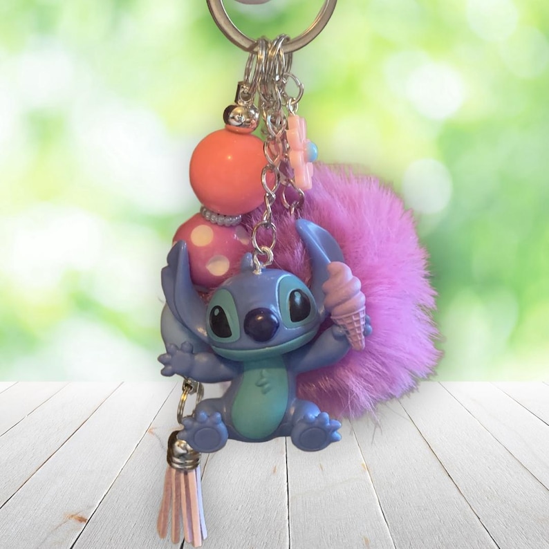 Puede incluir: Un llavero de Stitch azul y turquesa con un helado rosa, un pomp&oacute;n rosa y una borla rosa. El llavero tambi&eacute;n presenta una cuenta de lunares rosa y blanca y un dije de flor rosa.