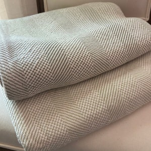 May include: Two folded blankets with a woven herringbone pattern. The blankets are in shades of light green and white, creating a textured appearance. The blankets are resting on a white surface.