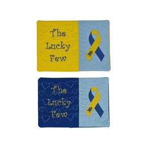 May include: Two rectangular fabric items featuring embroidered heart patterns. The top item is yellow and light blue with the text "The Lucky Few" and a yellow and blue ribbon. The bottom item is dark blue and light blue with the same text and ribbon.