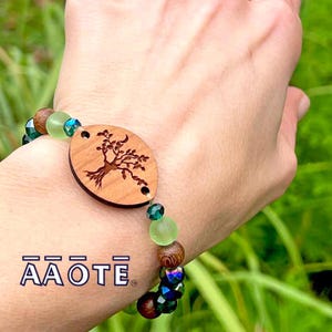 Puede incluir: Un brazalete de madera con un dije de árbol de la vida, cuentas de vidrio verde y azul y una cuenta de madera marrón. El brazalete está en la muñeca de una persona.