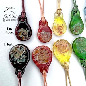 Könnte beinhalten: Sortiment an Glasanhängern mit wirbelnden Mustern in verschiedenen Farben, darunter Rot, Grün, Gelb und Schwarz. Die Anhänger sind an Schnüren befestigt und mit "Tiny Fidget" und "Fidget" beschriftet.