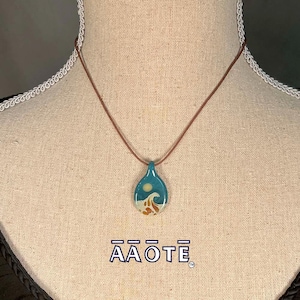 Könnte beinhalten: Eine braune Lederkordelkette mit einem türkisfarbenen Glasanhänger, der eine weiße Sonne und ein Wellendesign zeigt. Der Anhänger ist tropfenförmig. Die Kette wird an einer Puppe präsentiert. Der Text "AAŌTE" ist unterhalb der Kette aufgedruckt.