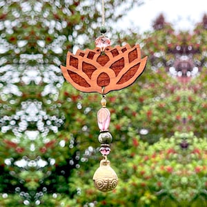 May include: A wooden lotus flower wind chime with pink and green glass beads and a gold bell. The chime is hanging from a string in front of a blurred background of green foliage.