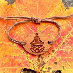 May include: A brown wooden bracelet with a circular charm featuring a silhouette of a person meditating on a lotus flower. The bracelet is made of brown cord and is tied with a knot.