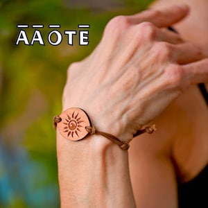 May include: A brown leather cord bracelet featuring a circular wooden charm engraved with a sun design. The charm displays a central spiral surrounded by radiating rays. The text AAOTE is visible in the upper left corner of the image.