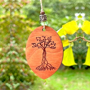 May include: A wooden oval ornament with a tree of life design engraved on it. The ornament is hanging from a string with a silver bead and a green glass bead.