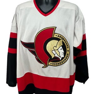 May include: White hockey jersey with black and red accents. Features a large team logo on the chest, with a red and gold emblem. The jersey has a V-neck collar and black sleeves with red stripes.