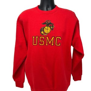 May include: A red crew neck sweatshirt featuring the USMC emblem and the letters "USMC" in yellow and black. The sweatshirt is displayed on a black mannequin.