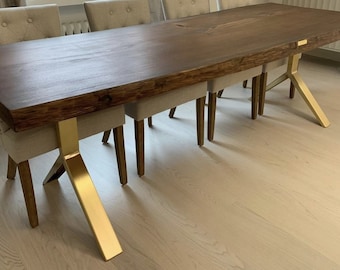 May include: A large, rectangular wooden dining table with a dark brown finish and a live edge. The table is supported by gold-colored metal legs. Beige upholstered chairs with wooden legs are arranged around the table. The table measures approximately 100 inches long.