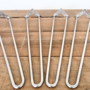 Könnte beinhalten: Vier silberne Haarnadel-Tischbeine. Jedes Bein hat eine dreieckige Montageplatte mit vorgebohrten Löchern. Die Beine sind aus Metall und haben ein gebogenes Unterteil. Ideal für DIY-Möbelprojekte. Jedes Bein ist etwa 76 cm lang.