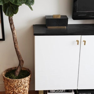 May include: A white cabinet with two doors and gold handles. The cabinet is sitting on gold legs and has a black top. There are books stacked underneath the cabinet, including "The Big Art", "In Flagrante Collecto", "Bright Bazaar", "Vogue", and "The Allure of the Women". There is a black box with a gold top on the cabinet. A large potted plant is to the left of the cabinet.