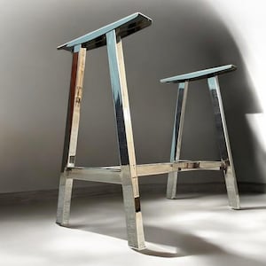 Op de afbeelding: Een tafelonderstel van verchroomd metaal met een eenvoudig, modern ontwerp. Het onderstel bestaat uit twee A-vormige frames die met een horizontale balk zijn verbonden.