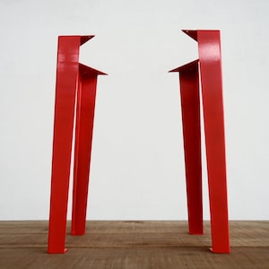May include: Two red metal table legs with a triangular design. The legs are standing on a wooden surface.