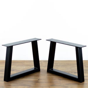 May include: Two black metal table legs with a modern, angled design. Each leg has a rectangular top and a trapezoidal frame, suitable for DIY furniture projects. The legs are shown against a white background.