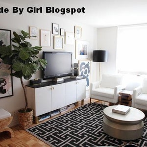 A living room interior with white walls, a white couch, a white coffee table, a white entertainment center with a flat screen TV, a large green plant in a woven basket, and a black and white geometric rug. The text 'Made By Girl Blogspot' is visible in the top left corner of the image.