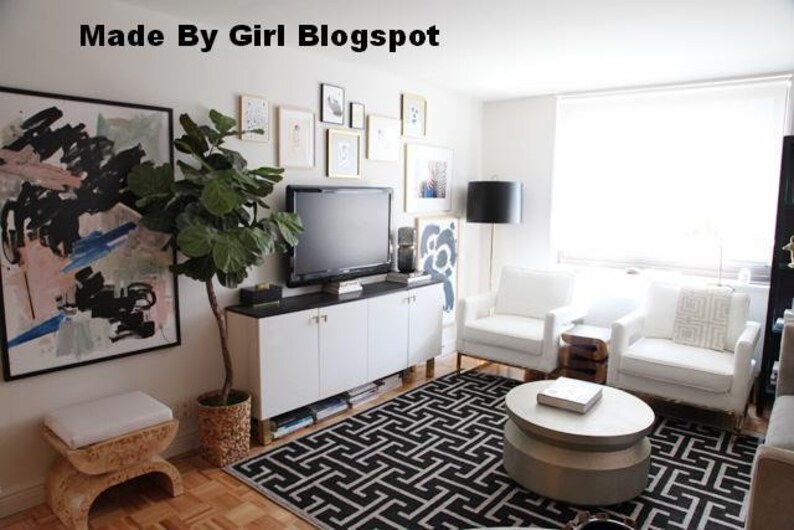 A living room interior with white walls, a white couch, a white coffee table, a white entertainment center with a flat screen TV, a large green plant in a woven basket, and a black and white geometric rug. The text 'Made By Girl Blogspot' is visible in the top left corner of the image.