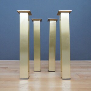 Four gold metal table legs with square tops. The legs are standing on a light wood floor.