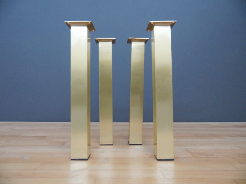 Four gold metal table legs with square tops. The legs are standing on a light wood floor.