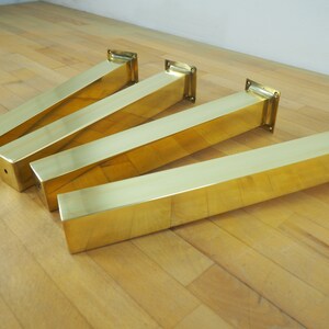 Four gold metal table legs with square bases. The legs are arranged in a row on a wood floor.
