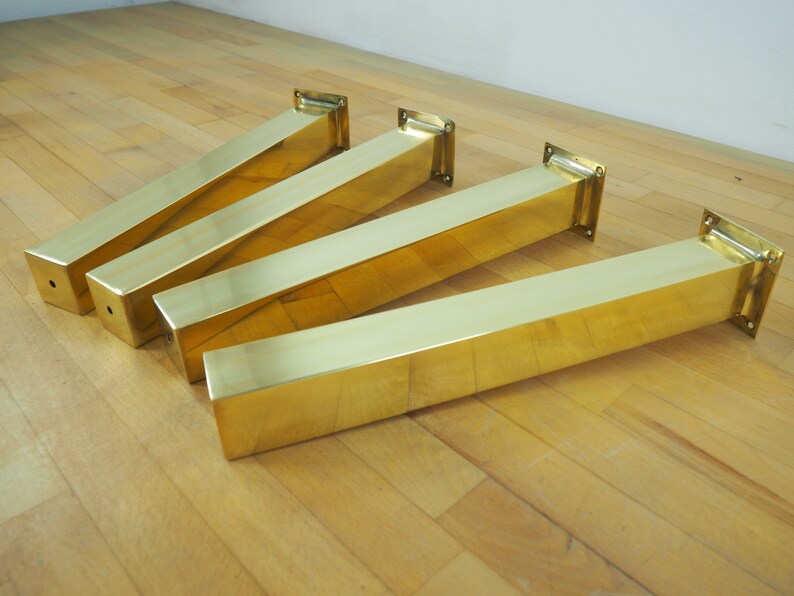 Four gold metal table legs with square bases. The legs are arranged in a row on a wood floor.