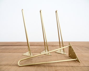 May include: Four gold metal hairpin legs for furniture. The legs are angled and have a triangular base. The legs are arranged in a group, with one leg lying on top of the others.