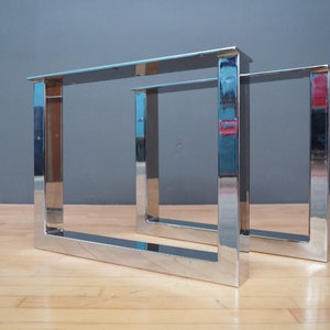 May include: Two silver metal table legs in a square shape. The legs are made of polished metal and have a reflective surface.