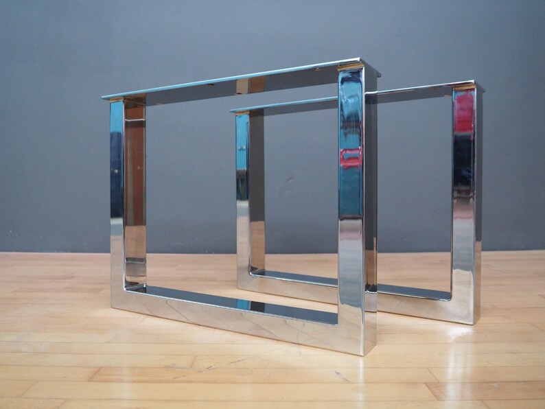 May include: Two silver metal table legs in a square shape. The legs are made of polished metal and have a reflective surface.