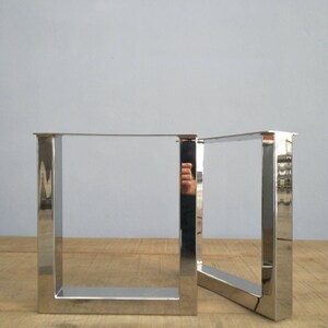 May include: Two silver metal table legs in a square shape. The legs are made of polished metal and have a modern design.