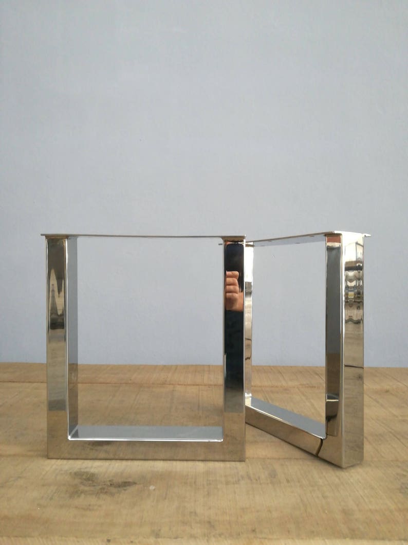 May include: Two silver metal table legs in a square shape. The legs are made of polished metal and have a modern design.