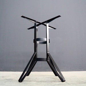 Op de afbeelding: Zwarte metalen tafelpoot met een kruisvormige bovenkant en drie poten. De poot is ontworpen om een tafelblad te dragen.