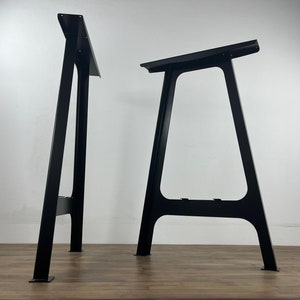 Two black metal table legs, each with a triangular shape and a flat top. The legs are standing on a wooden surface.