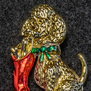 Op de afbeelding: Een gouden hondenfiguur dat een rode sok vasthoudt met een groene strik. Het figuur is een kerstversiering.