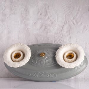 May include: A vintage-style ceiling light fixture with two white, floral-edged bulb holders. The gray base features ornate detailing and a gold-colored center accent. The light fixture is designed for overhead illumination.