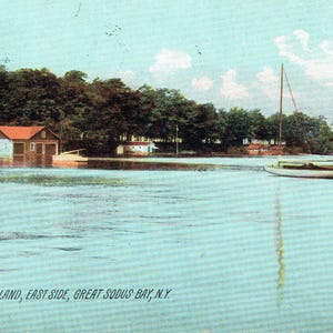 May include: A vintage postcard depicting a tranquil waterscape with a sailboat and a boathouse. The boathouse has a red roof and is surrounded by green trees. The text at the bottom reads "Isley Island, East Side, Great Sodus Bay, N.Y."